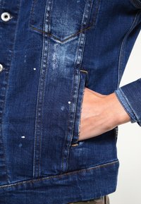 Jeansjacka i mörkblått med en strukturerad yta. Har knäppning med knapp, framfickor och slitna områden med vita färgstänk.