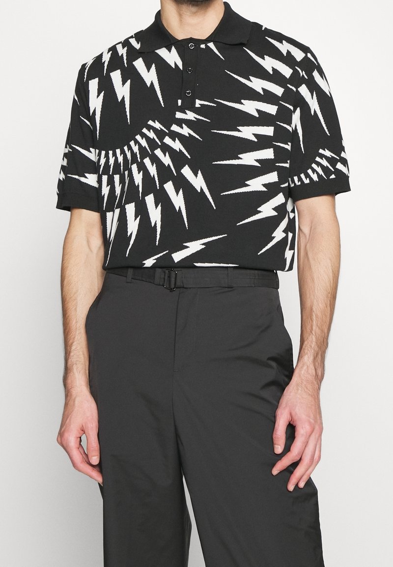Homme portant un polo noir avec un motif d'éclair blanc et un pantalon noir, debout devant un fond uni.