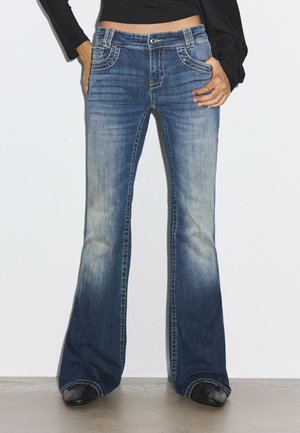 Femme portant un jean bootcut bleu délavé, les mains dans les poches et des chaussures noires, debout devant un fond clair uni.