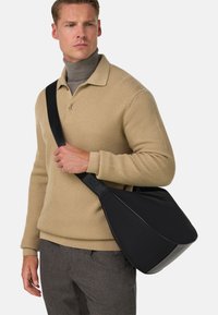 Borsa a spalla nera realizzata in materiale liscio con un design sagomato; presenta una tracolla ampia e dettagli sottili. Indossata sopra un maglione beige.