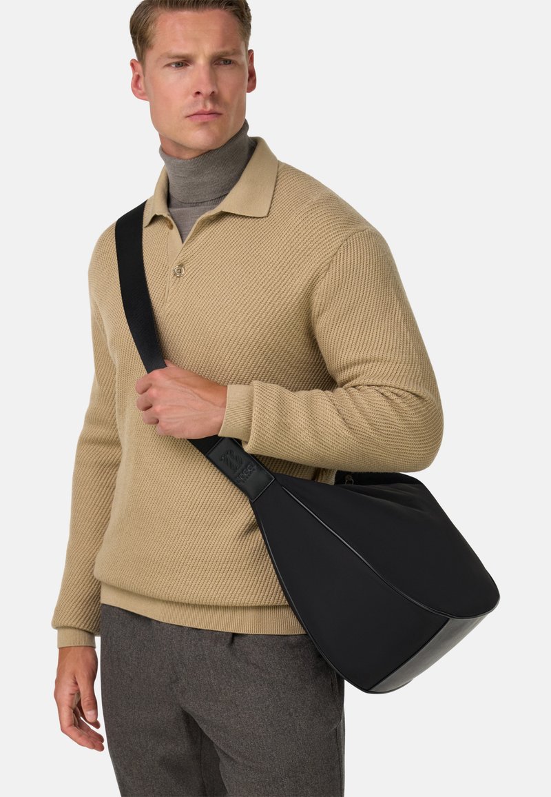 Borsa a spalla nera realizzata in materiale liscio con un design sagomato; presenta una tracolla ampia e dettagli sottili. Indossata sopra un maglione beige.