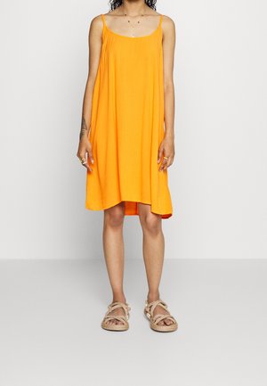 Vrouw draagt een losvallende, mouwloze oranje jurk met dunne bandjes en beige sandaalriemen, staand tegen een effen lichte achtergrond.