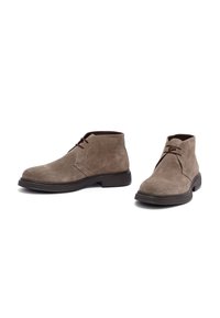 Par de botas de ante taupe hasta el tobillo con suelas y cordones marrón oscuro, exhibidas sobre un fondo blanco.