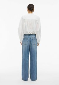 Blouse blanche à manches longues avec un détail plissé au dos, associée à un jean en denim bleu clair, ample, avec des poches arrière classiques.