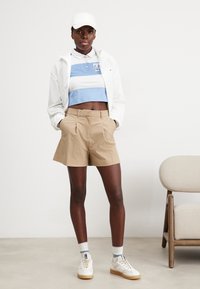 Chaqueta blanca, polo corto de rayas azules y blancas, pantalones cortos color beige, zapatillas blancas con acentos beige y gorra blanca. Tejido texturizado y líneas limpias.