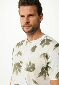 Vit t-shirt med grönt lövmönster, korta ärmar och rundad halsringning. Tillverkad av ett lätt tyg med en subtil textur.