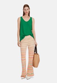Green sleeveless top, patterned wide-leg pants in yellow, orange, and pink zigzags, holding a tan handbag with dark handles, neutral shoes.