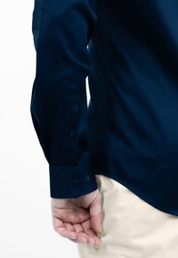 Marineblauw langemouwen shirt met knoopmanchetten, voorzien van een logo op de mouw. Soepele structuur en op maat gemaakte pasvorm. Lichtbeige broek op de achtergrond.