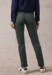 Pantalones ajustados de color verde oliva con bolsillos traseros, combinados con zapatillas blancas con detalles en negro. El modelo muestra un perfil lateral.