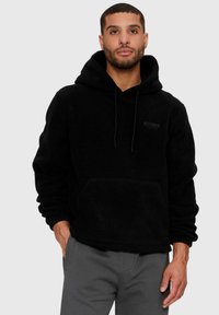 Mann in schwarzem Fleece-Hoody mit Kängurutasche und grauen Hosen, der mit einer Hand in der Tasche vor einem schlichten Hintergrund steht.