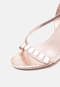 Πέδιλο με μεσαίο τακούνι σε ροζ χρυσό, με λουράκι σε σχήμα Τ και στρογγυλές μεταλλικές διακοσμήσεις κατά μήκος του μπροστινού λουριού.