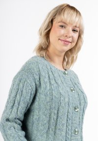 Ljusblågrön stickad cardigan med en texturerad kabelmönster, som har stora gröna knappar framtill och ribbade ärmslut.