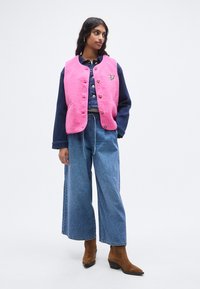 Ροζ γιλέκο από φλις με κλείσιμο με κουμπιά και λεπτομέρεια καρδιάς, φορεμένο πάνω από μπλούζα από τζιν και μπλε τζιν παντελόνι φαρδιάς γραμμής, ολοκληρωμένο με καφέ μποτάκια μέχρι τον αστράγαλο.