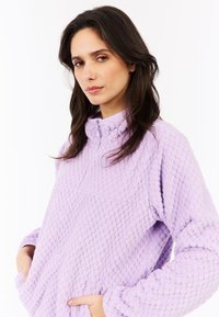 Žena s dlhými tmavými vlasmi, oblečená v textúrovanom svetri levanduľovej farby s golierom a bočnými vreckami, s rukami zasunutými vo vreckách, s neutrálnym výrazom.