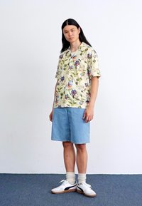 Blomstret kortærmet skjorte med gul baggrund, parret med blå denimshorts. Model iført hvide sneakers med sorte detaljer.