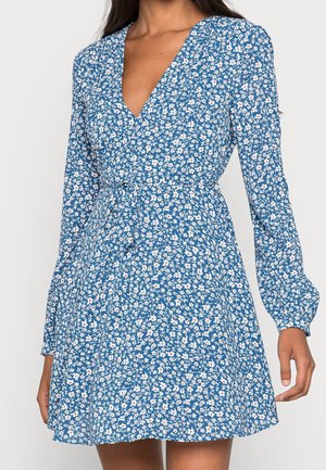 Femme portant une robe bleue à manches longues avec un petit motif floral blanc et rose et un décolleté en V, cintrée à la taille avec une ceinture en tissu.