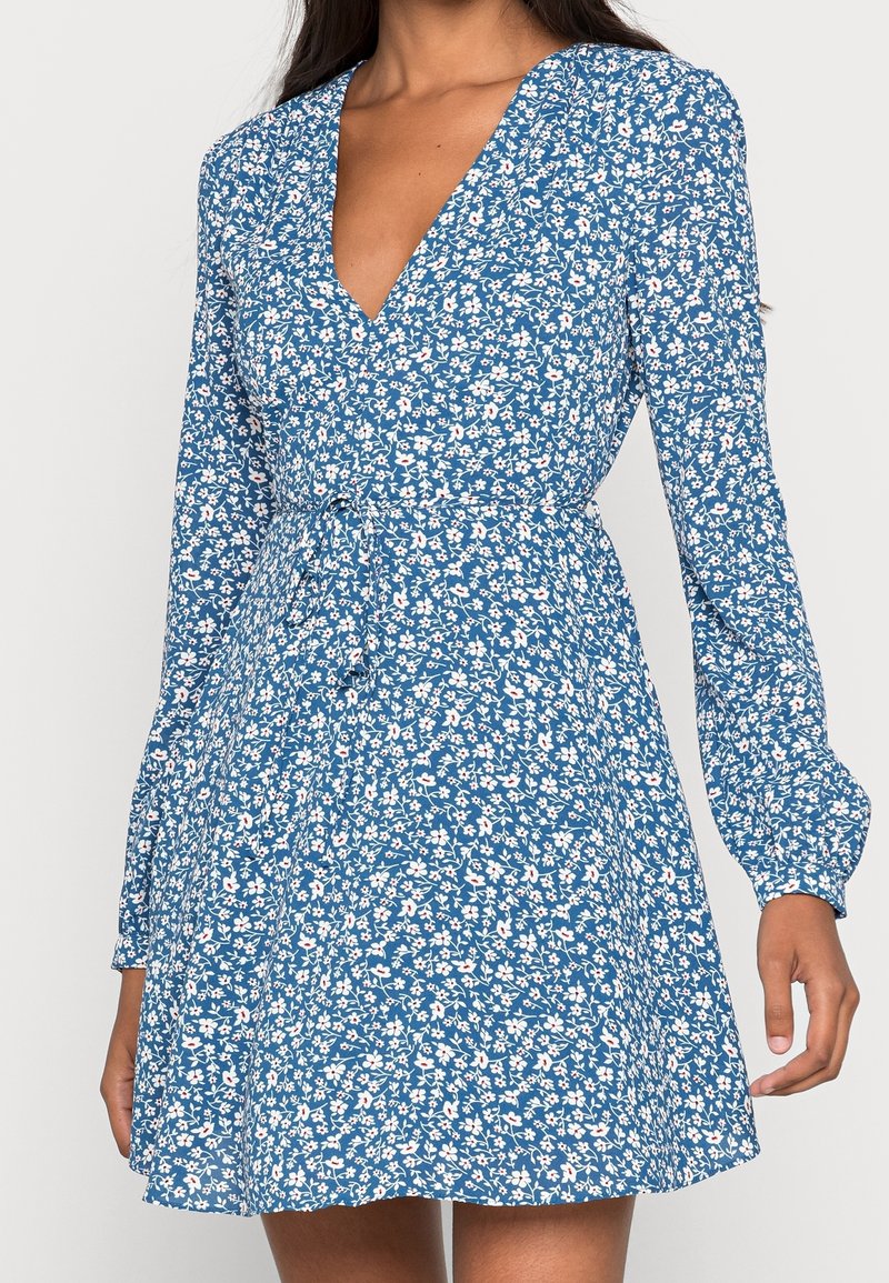Femme portant une robe bleue à manches longues avec un petit motif floral blanc et rose et un décolleté en V, cintrée à la taille avec une ceinture en tissu.