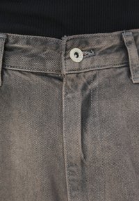 Acercamiento al cinturón de unos jeans de mezclilla gris con botón de metal y trabillas para el cinturón, combinados con una blusa negra de canalé.