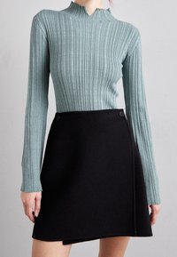 Žebrovaný tyrkysový top s dlouhým rukávem a vysokým límcem, doplněný černou zavinovací sukní se bočními knoflíky, hladkou texturou a přiléhavým střihem.