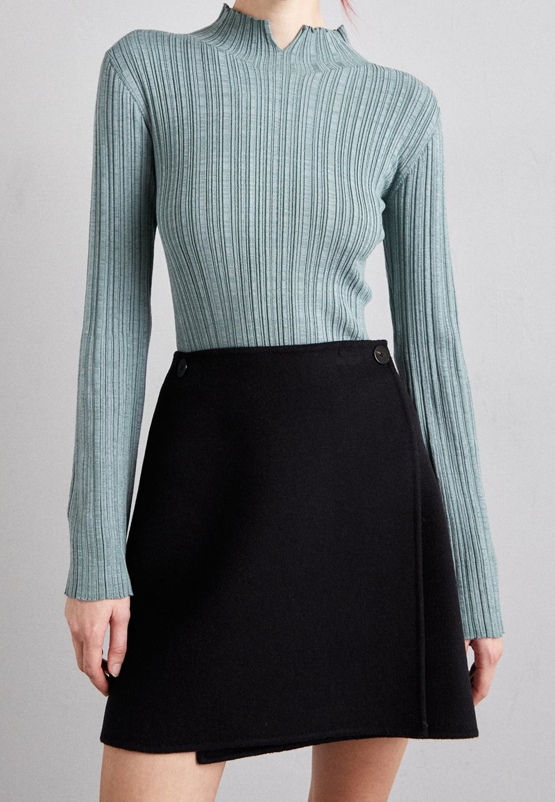 Žebrovaný tyrkysový top s dlouhým rukávem a vysokým límcem, doplněný černou zavinovací sukní se bočními knoflíky, hladkou texturou a přil�éhavým střihem.