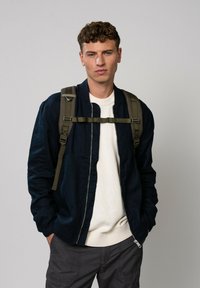 Veste de bombardier marine avec fermeture éclair, portée sur un pull crème. Associée à un sac à dos vert olive, le mannequin porte un pantalon cargo gris.