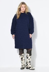 Marineblaumes, gestepptes Kleid mit langen Ärmeln und lässigem Schnitt, kombiniert mit kniehohen Stiefeln im Kuhmuster. Glatte Textur und ausgeprägtes Muster.