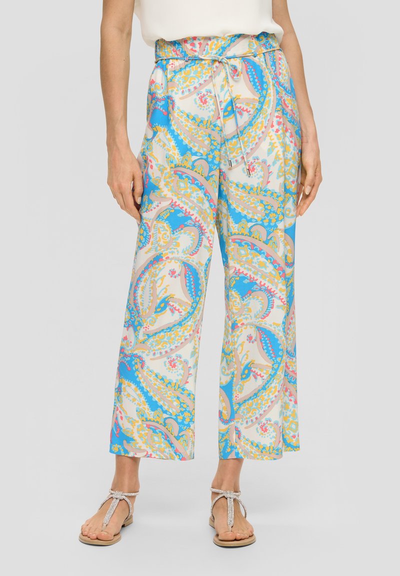 Femme portant un pantalon à jambes larges avec un motif cachemire bleu, jaune et rose, taille nouée, associé à des sandales beige et un haut blanc.