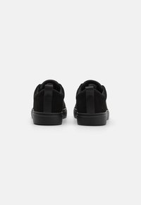 Ett par svarta låga sneakers med dragflikar på hälarna, sedda från baksidan mot en enkel vit bakgrund.