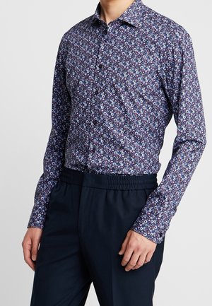 Mand iført en langærmet skjorte med små blomsterprint i nuancer af blå, hvid og lilla, kombineret med mørke marineblå bukser.