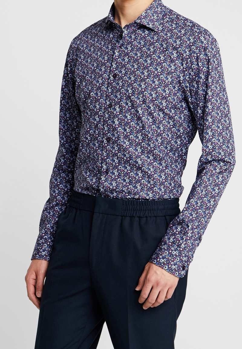 Homme portant une chemise à manches longues boutonnée avec un petit motif floral dans des tons de bleu, blanc et violet, associée à un pantalon bleu marine foncé.