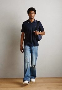 Ναυτικό πόλο μπλουζάκι με λευκό γιακά, ανοιχτό μπλε φθαρμένα τζιν και λευκά αθλητικά παπούτσια. Κρατώντας μια μικρή ναυτική τσάντα τον ώμο. Ξύλινο πάτωμα στο φόντο.