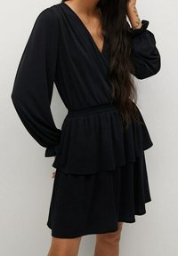 Vestido negro de envoltura con mangas largas, escote en v, dobladillo con volantes en capas y cintura elástica. Fabricado en un tejido suave y fluido.