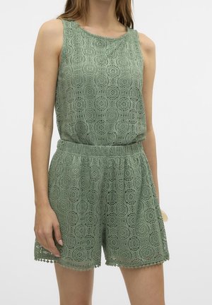 Kobieta w zielonym, bezrękawnym topie z koronkowej dzianiny i dopasowanych szortach z elastycznym paskiem na tle jednolitego tła.