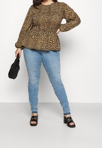Frau trägt ein braunes Leopardenmuster-Langarmoberteil, helle blaue Skinny-Jeans, schwarze hochhackige Sandalen und hält eine kleine schwarze Handtasche.