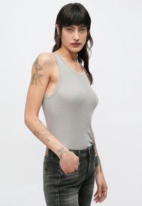 Žena s tamnom kosom, odjevena u sivu majicu na bretele s rebrastim uzorkom i crne traperice, pokazuje tetovaže na lijevoj ruci i ruci u opuštenom položaju.