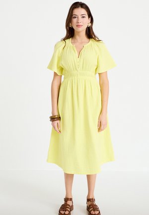 Vrouw draagt een gele jurk tot op de knie met korte mouwen en een geplooide taille, bruine sandalen, bloemsieraden en houten armbanden om haar pols.