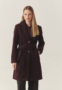 Burgundská vlněná kabátová směs s opaskem v pase, s výstřihy a dvěma knoflíky. Dlouhé rukávy a boční kapsy dotvářejí design.