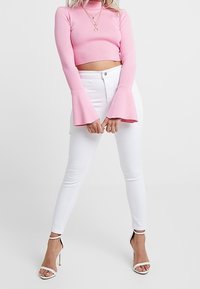 Růžový žebrovaný croptop s dlouhými, rozšířenými rukávy, spárovaný s upnutými bílými džíny a lodičkami na kotníkovém popruhu. Jednoduchý vrstvený náhrdelník podtrhuje celkový vzhled.