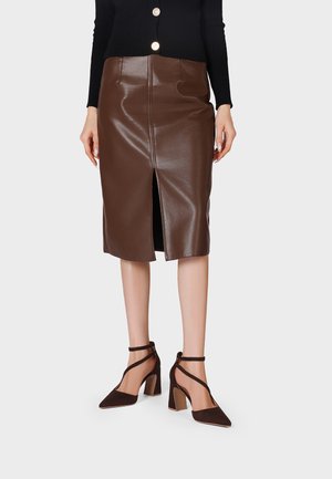 Donna che indossa una gonna a matita di pelle marrone al ginocchio con spacco frontale e scarpe con tacchi alti marrone scuro con cinturini su sfondo bianco.