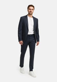 Tamno plavi mornarski odijelo s jednorednim sakoom i odgovarajućim hlačama. Nošeno preko bijele majice i upareno s bijelim tenisicama. Glatka tekstura.