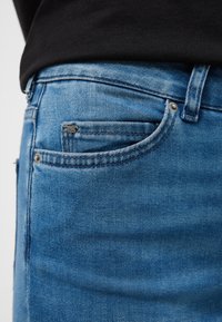 Gaiši zili džinsa džinsi ar standarta piecu kabatu dizainu, kontrastējošām šuvēm un metāla akcentu priekšējā kabatā.
