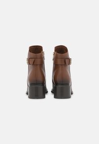 Botas de tobillo de cuero marrón con un tacón cuadrado corto, que cuentan con una cremallera lateral, detalle de hebilla y una textura suave.