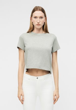 Γυναίκα με ίσια ανοιχτό καστανά μαλλιά που φοράει κρίκους στα αυτιά, γκρι κοντομάνικη μπλούζα και λευκά παντελόνια ψηλόμεσα, μπροστά σε απλό φόντο.