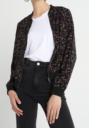 Vrouw draagt een zwart bloemenprint vest met rits over een wit T-shirt en hoge zwarte jeans tegen een effen achtergrond.