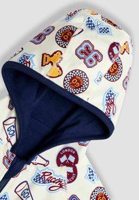 Hanorac din bumbac, cu glugă, de culoare crem, cu grafică vibrantă inspirată din curse. Accente de culoare albastru marin de-a lungul fermoarului și pe margini.