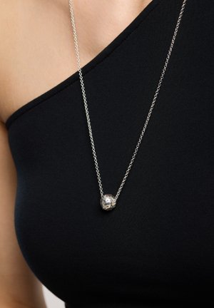 Collar de cadena de plata que presenta un colgante redondo y con patrón, con una piedra central, llevado sobre una blusa negra de un solo hombro.