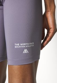Fioletowe spodenki sportowe z elastycznego materiału, z białym logo i napisem "THE NORTH FACE MOUNTAIN ATHLETICS" z boku.