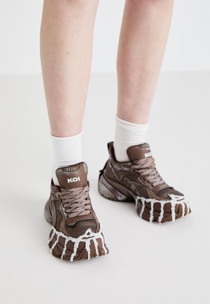 Pruunid võrkmaterjalist spordijalatsid, millel on paksud tallad valge tilkuva kujundusega, tekstuuriga kingapaelad ja logoga kontsakaunistused. Üksikasjad on valged sokid, mis paistavad üle ääre.