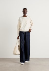 Felpa crema con motivo testurizzato sul colletto, abbinata a jeans larghi scuri. La modella tiene una borsa beige e si trova su un pavimento chiaro.