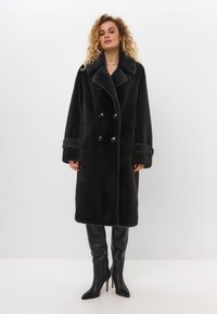 Schwarzer Kunstfell-Mantel mit doppelter Knopfleiste, weitem Kragen, Knopfdetails und weitem Ärmeln. Das Modell trägt schwarze, bis zum Oberschenkel reichende Stiletto-Stiefel.